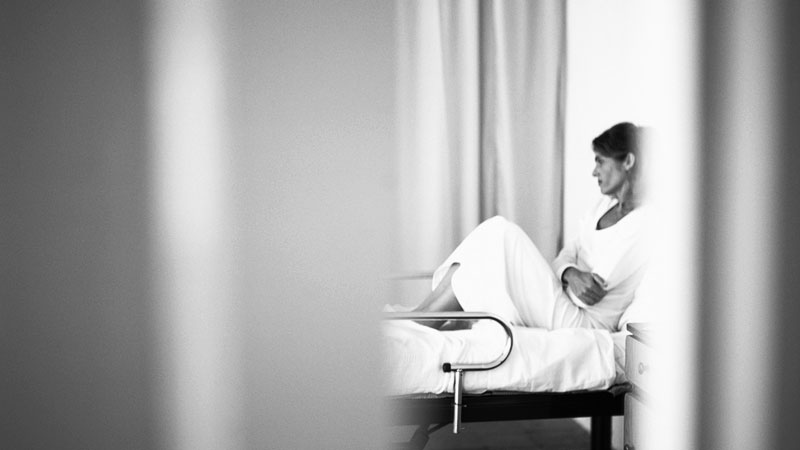 Blick durch einen Vorhang auf ein Krankenbett mit einer Frau, die ihre Arme verschränkt.